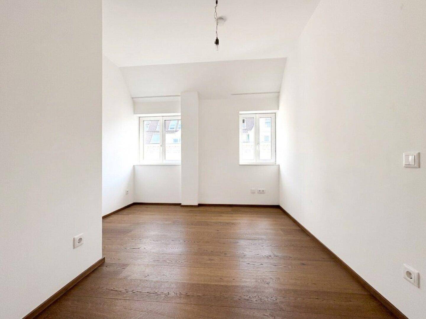 Helles, leeres Zimmer mit Holzboden und zwei Fenstern, vielseitig nutzbar als Schlafzimmer oder Arbeitszimmer.