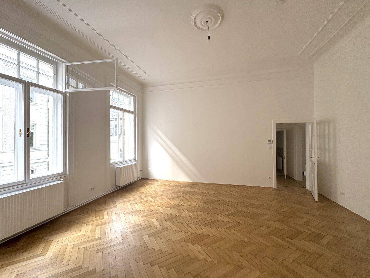 Helles Wohnzimmer mit Fischgrätparkett, hohen Decken und großen Fenstern, die den Raum erhellen.