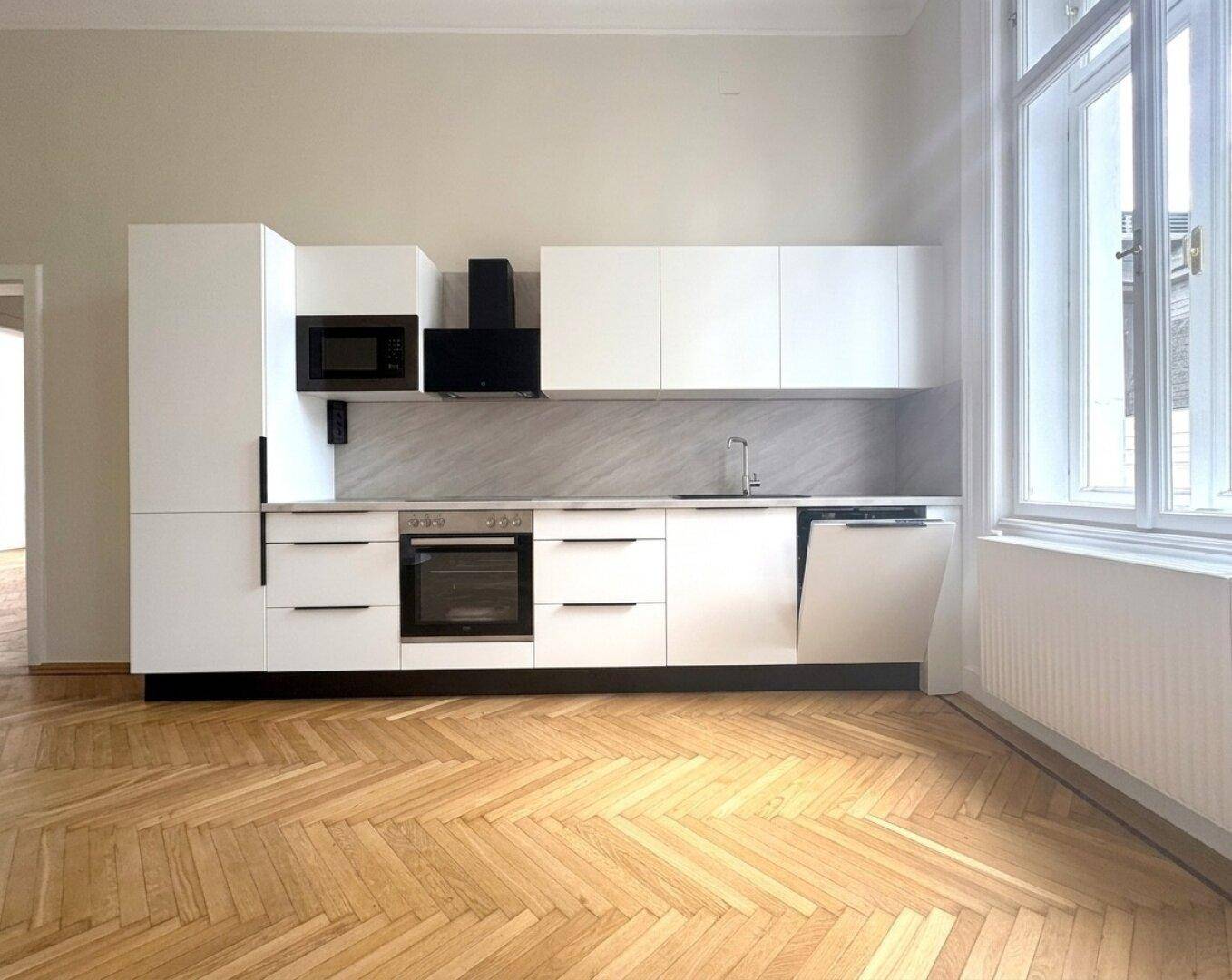 Moderne, weiße Einbauküche mit integrierten Geräten und Fischgrätparkett in einem hellen Raum.