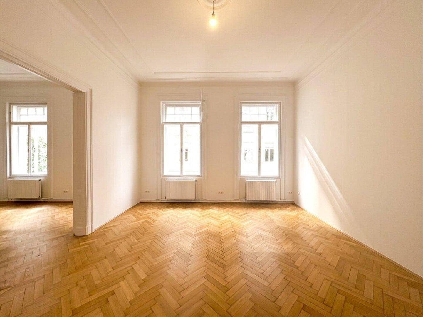 Heller Wohnraum mit Fischgrätparkett, hohen Decken und großen Fenstern, die viel Licht hereinlassen.