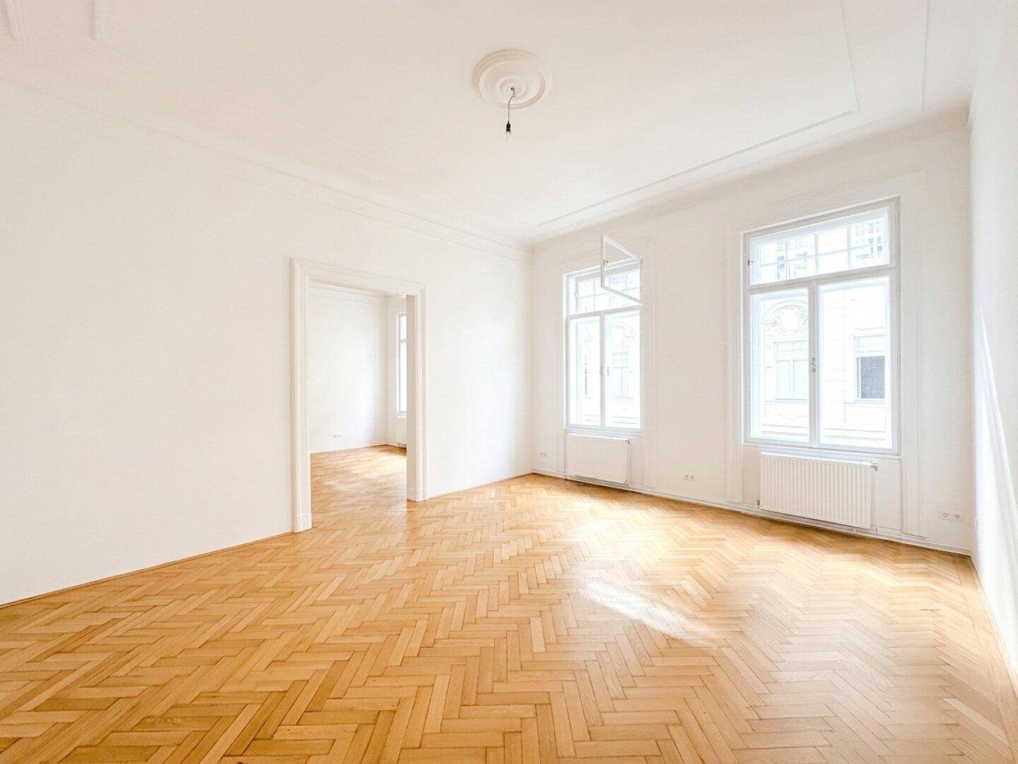 Geräumiges Wohnzimmer mit Fischgrätparkett, hohen Decken und großen Fenstern, die den Raum erhellen.