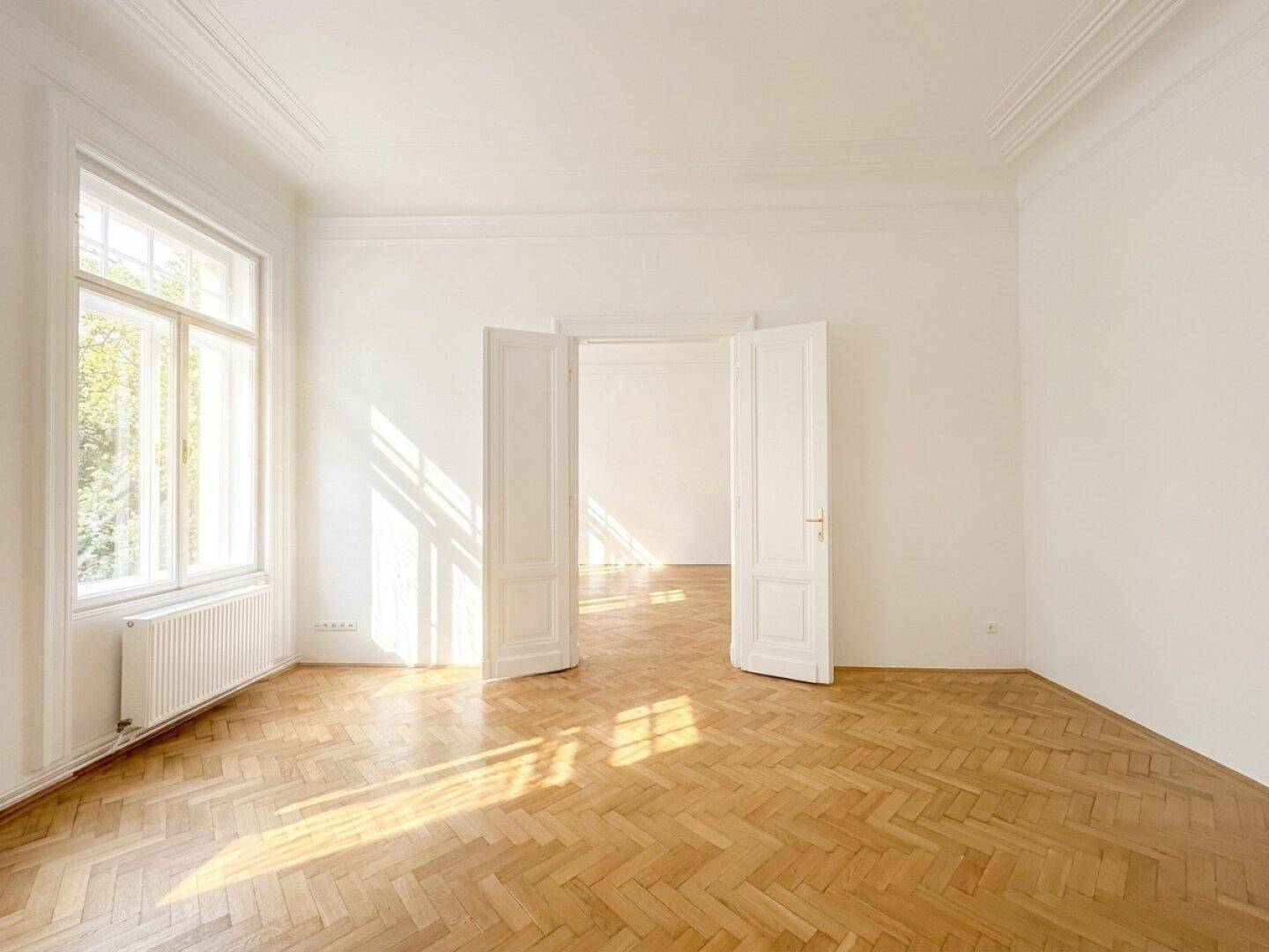 Lichtdurchflutetes Zimmer mit Fischgrätparkett und hohen Doppeltüren, die zu einem weiteren Raum führen.