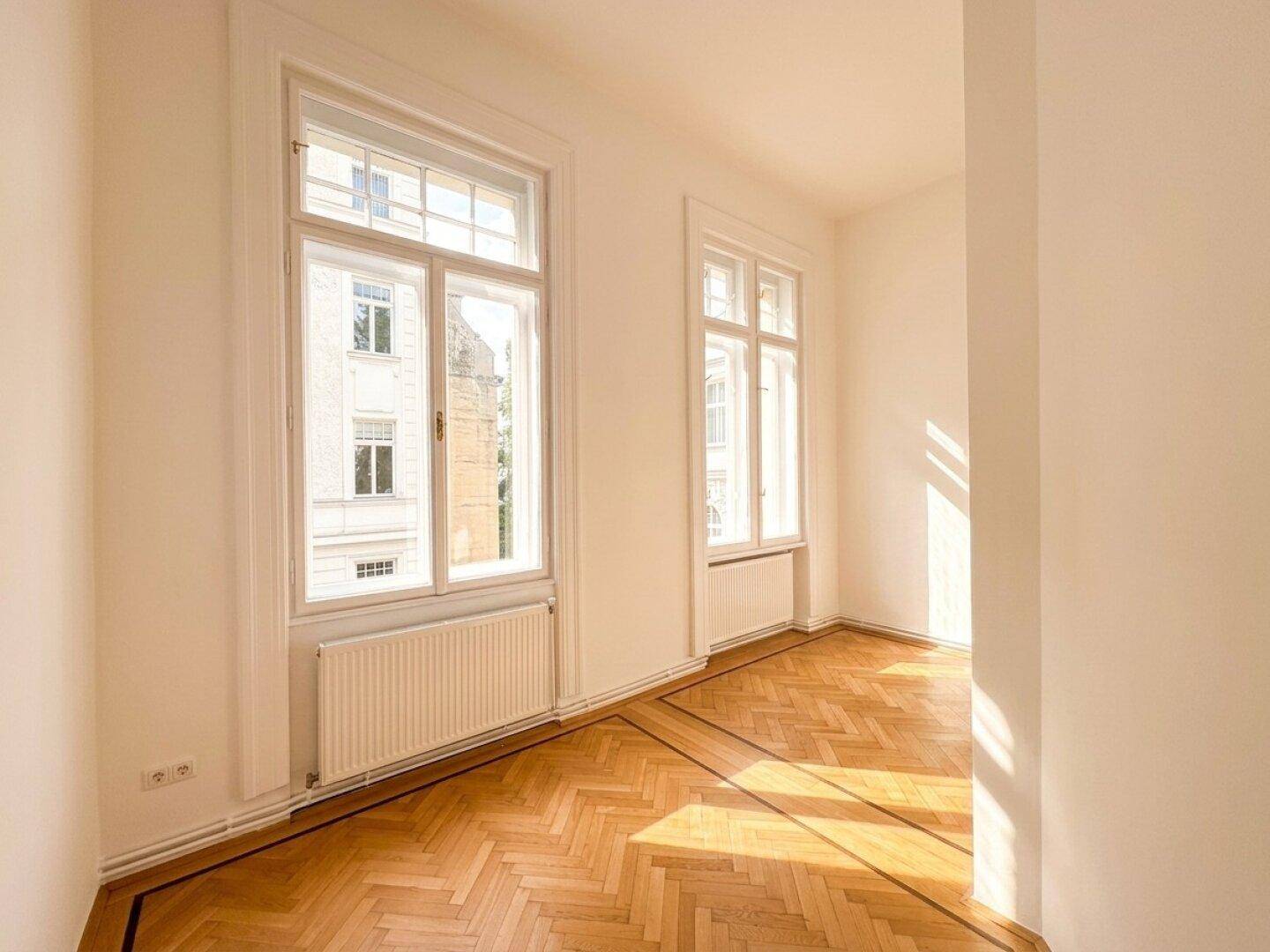 Helles Zimmer mit Fischgrätparkett und großen Fenstern, die viel Sonnenlicht hereinlassen.