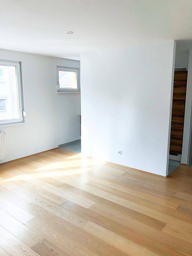 Heller Wohnbereich mit Holzparkettboden und mehreren Fenstern, die den Raum mit Tageslicht füllen.