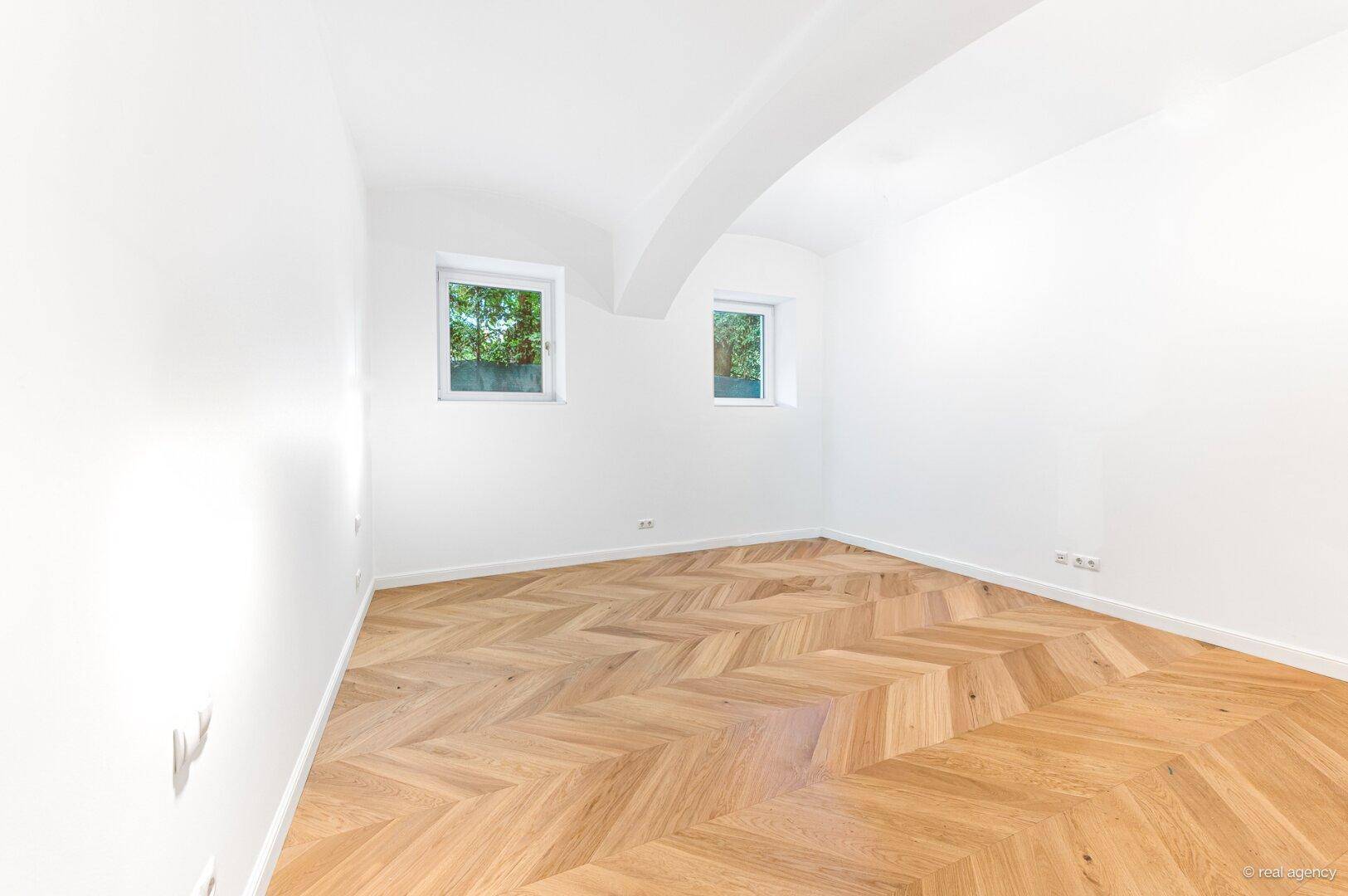 Ruhiges Zimmer mit Fischgrätparkett, weißen Wänden und zwei Fenstern, ideal als Schlafzimmer.