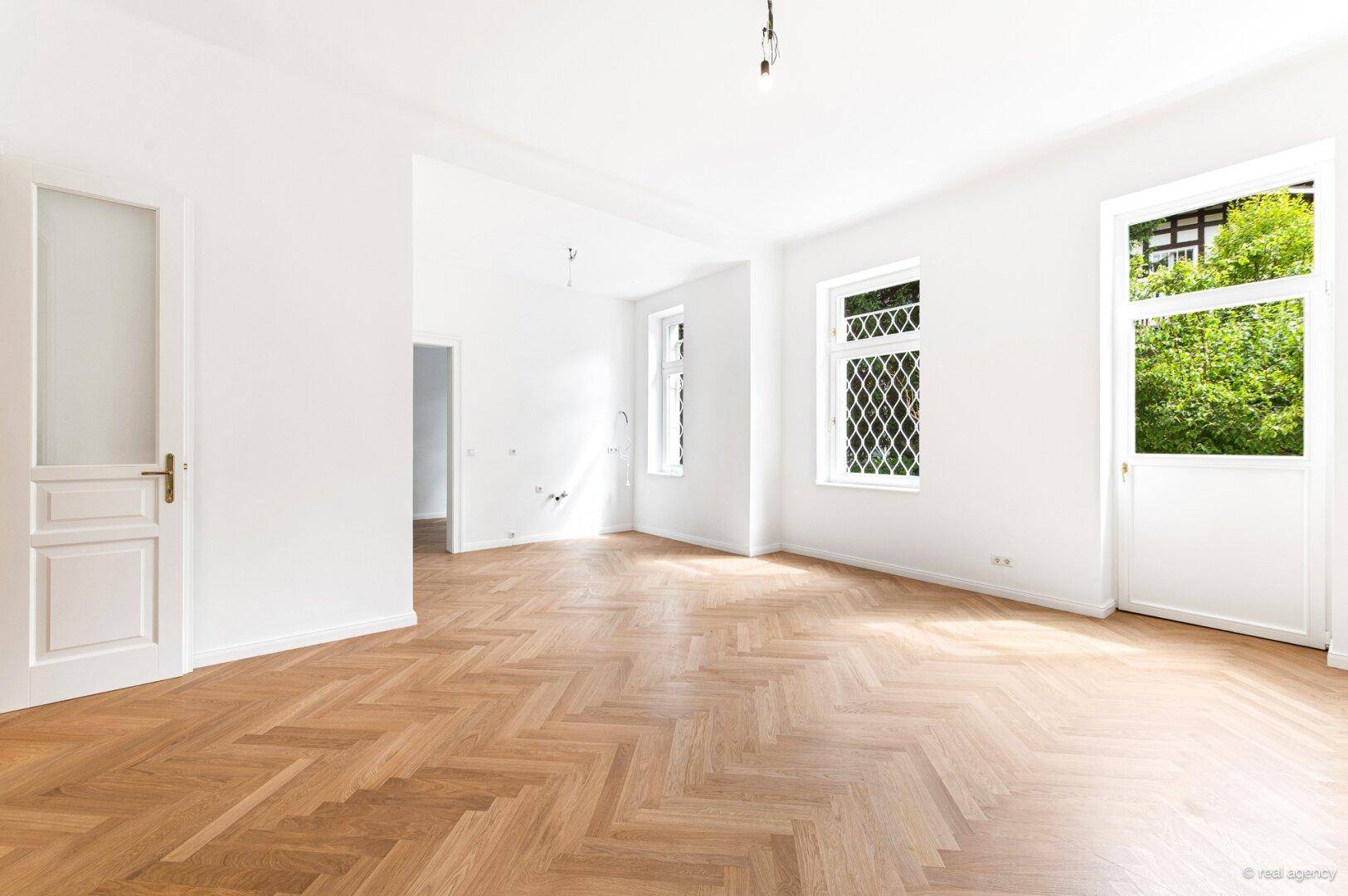 Helles, leeres Zimmer mit Fischgrätparkett und großen Fenstern mit Blick ins Grüne.