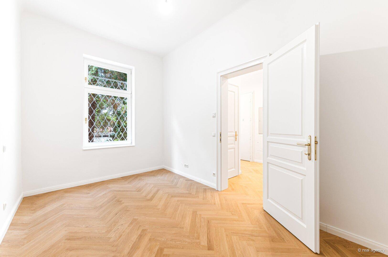 Helles Zimmer mit Fischgrätparkett und weißer Tür, bereit zur Einrichtung.