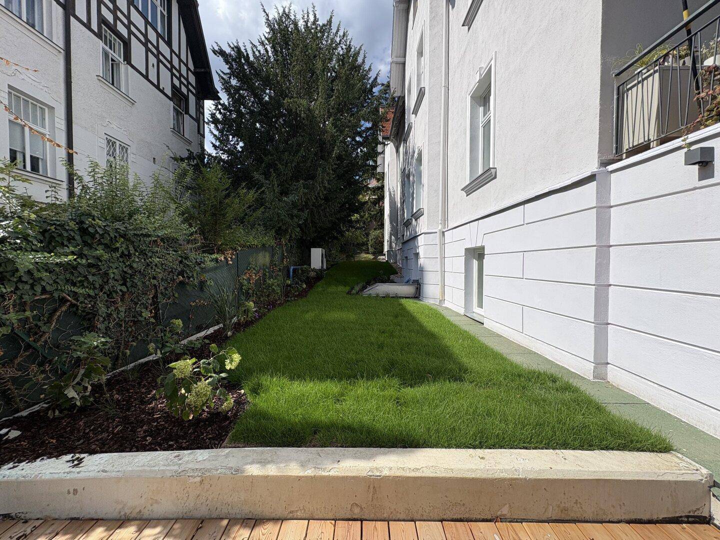 Schmaler Gartenbereich mit Rasenfläche und angrenzender Holzterrasse neben dem Gebäude.