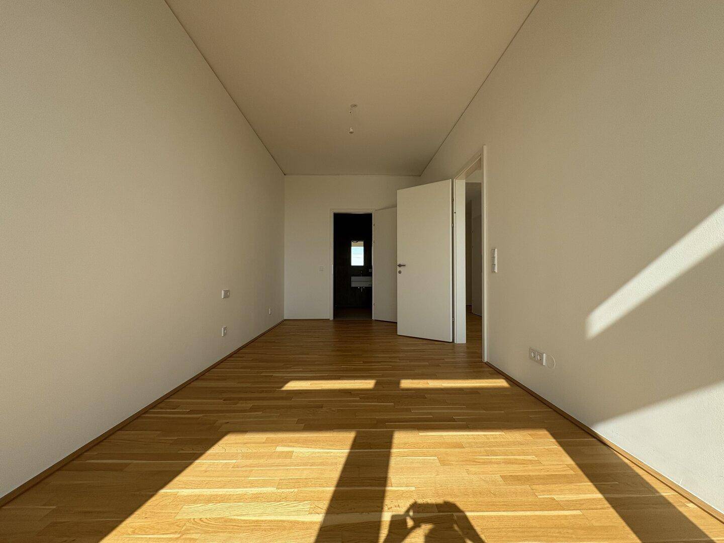 Leerer Raum mit hellem Holzboden und weißen Wänden, durchflutet von natürlichem Sonnenlicht.