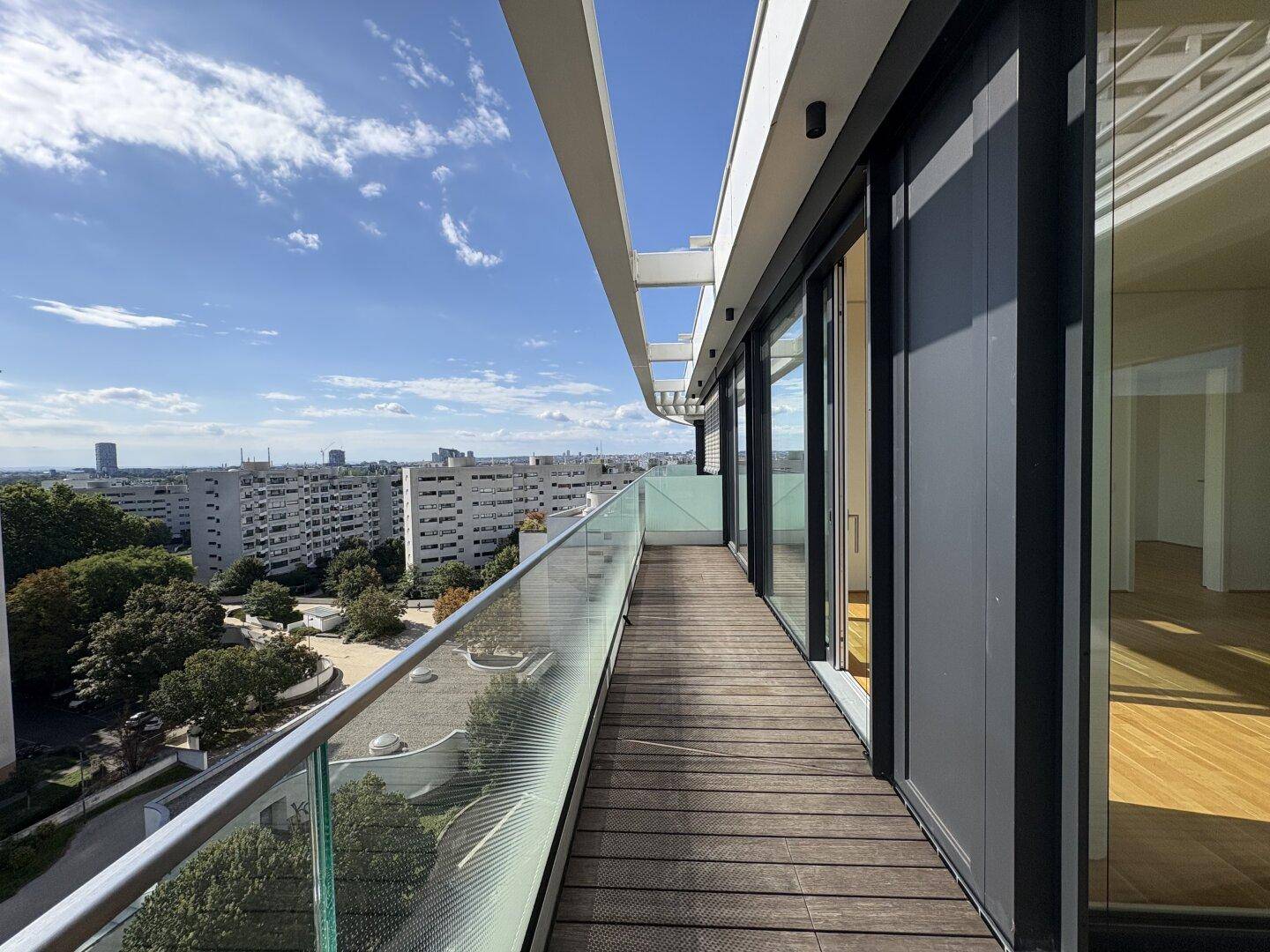 Langer Balkon mit Holzdeck und Glasgeländer, der einen weiten Panoramablick auf die Stadt und den See bietet.