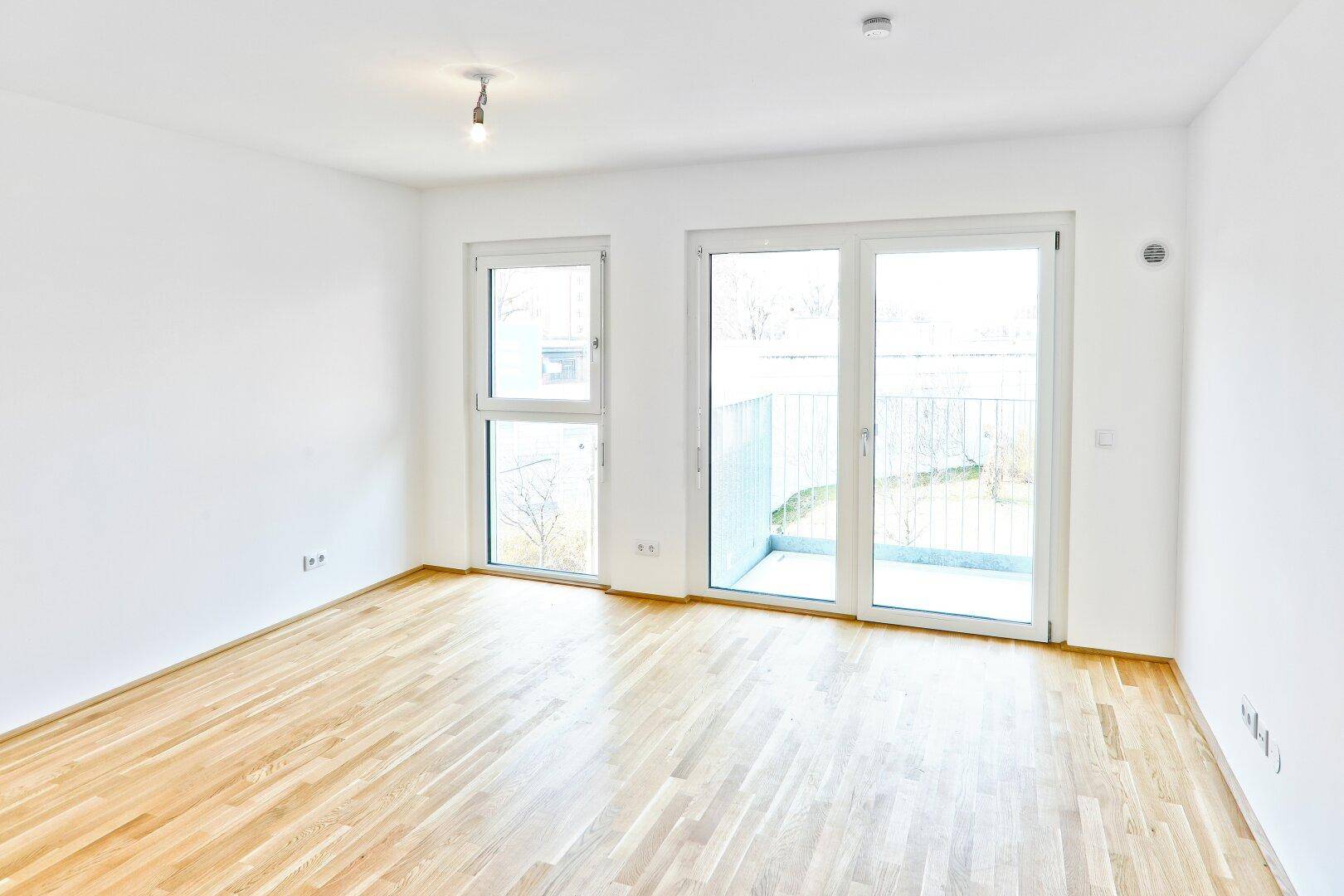 Geräumiges Zimmer mit hellem Parkettboden und großen Fenstern zum Balkon.