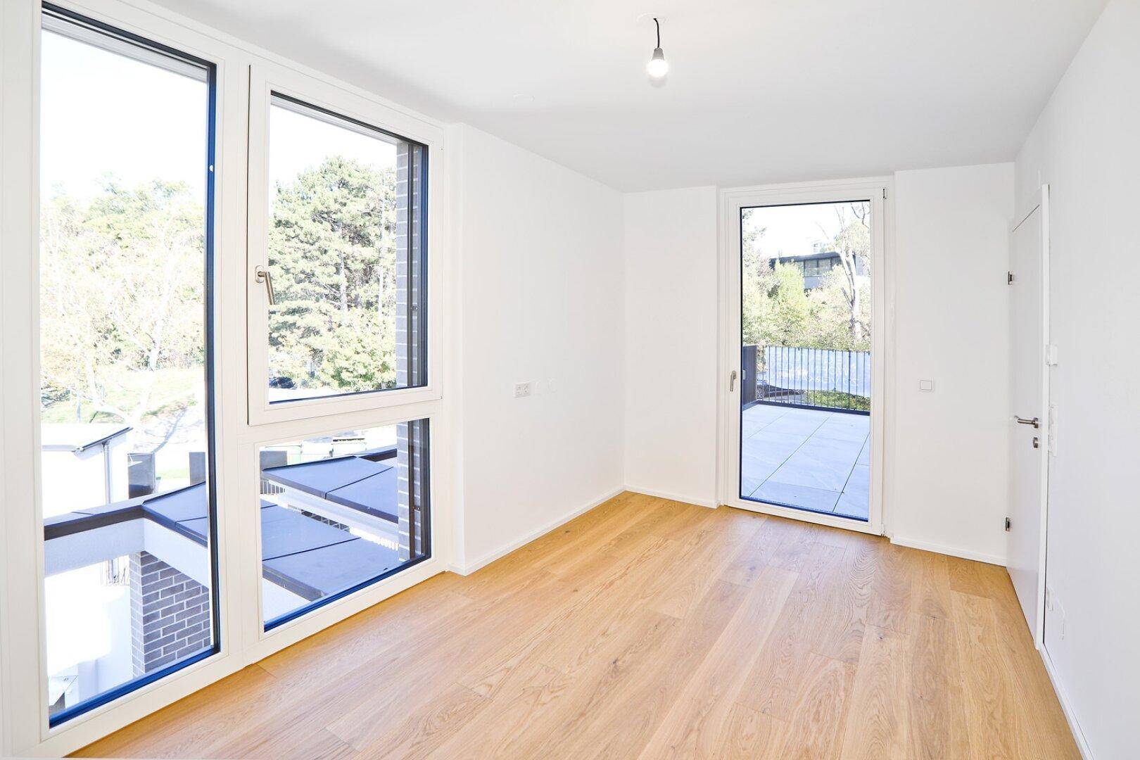 Helles Zimmer mit großen Fenstern und Zugang zu einem Balkon oder einer Terrasse mit Grünblick.