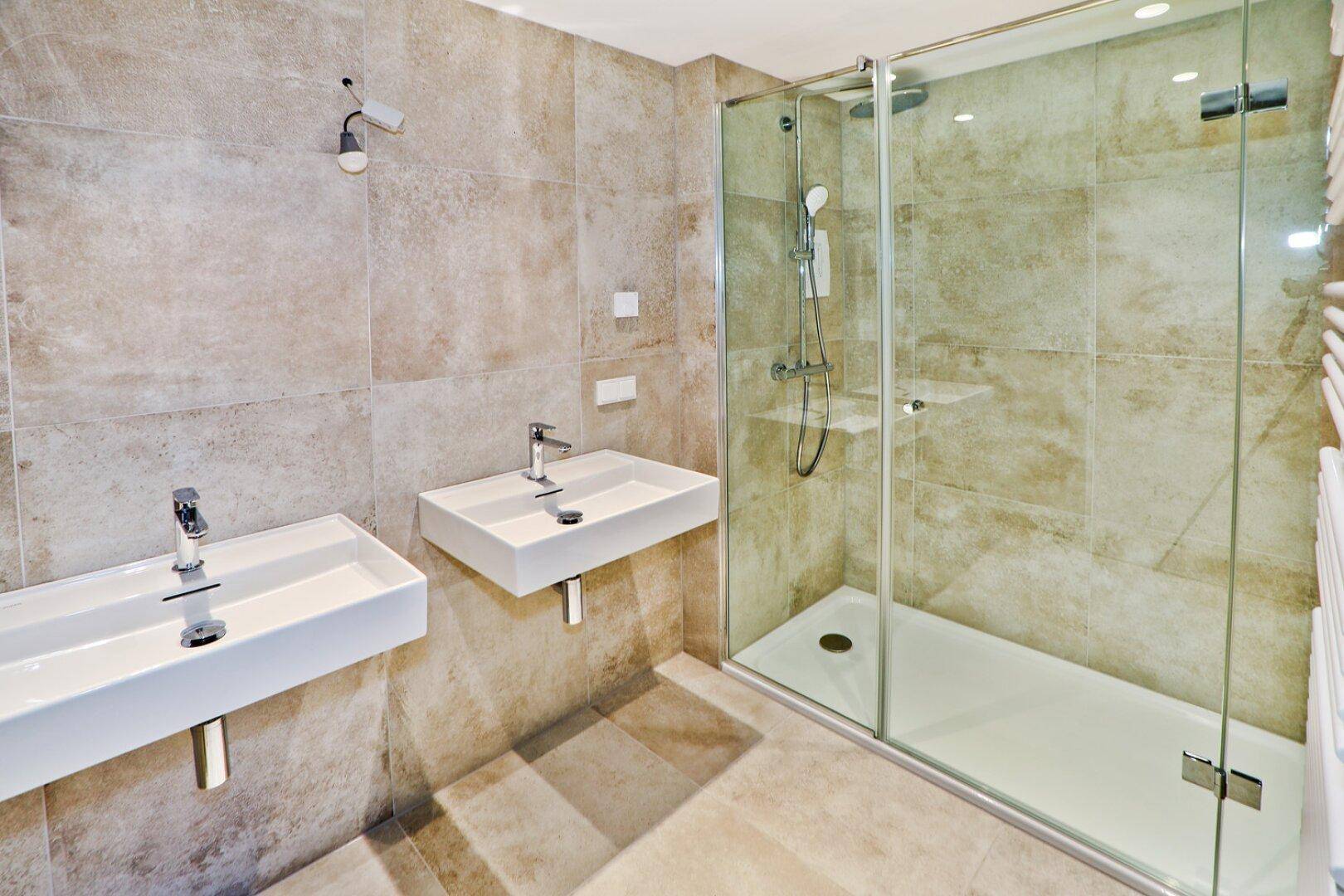 Modernes Badezimmer mit zwei Waschbecken, großer Duschkabine und beigefarbenen Fliesen.