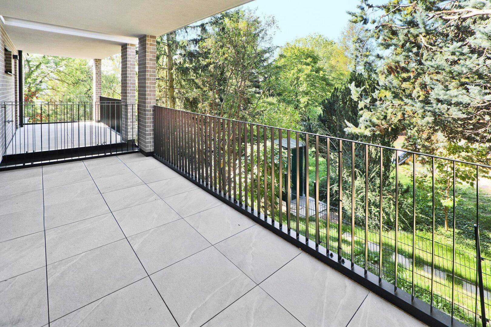 Heller Balkon mit Fliesenboden und Metallgeländer, umgeben von üppigem Grün.