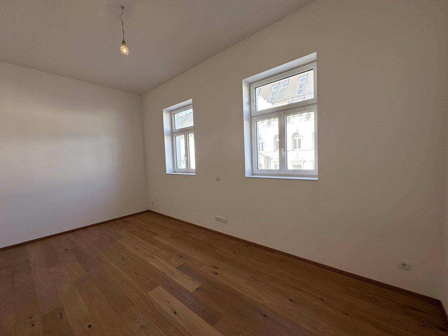 Leerer Raum mit hellem Holzfußboden und zwei Fenstern, die viel natürliches Licht hereinlassen.