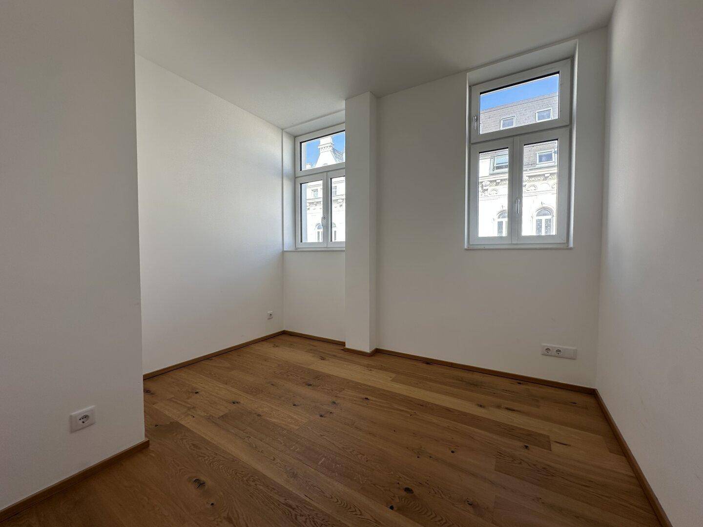 Helles Zimmer mit zwei Fenstern und Parkettboden, bietet viel Tageslicht und eine angenehme Atmosphäre.