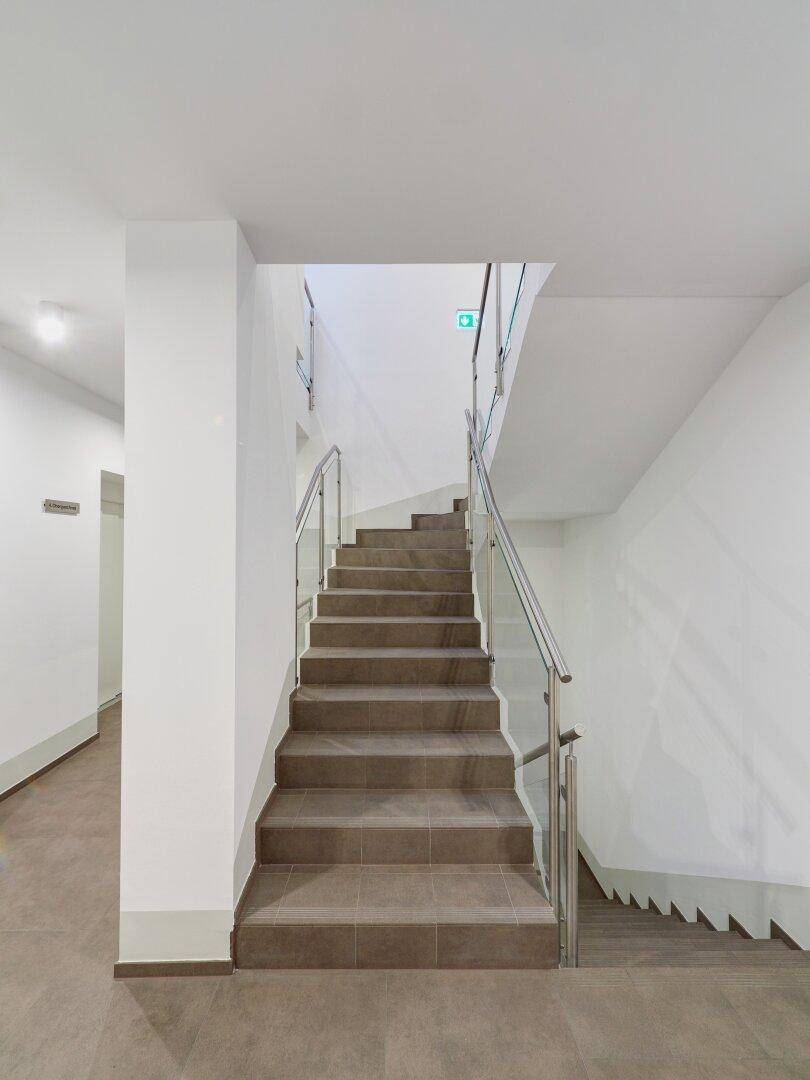 Geräumiges Treppenhaus mit gefliesten Stufen und modernem Geländer, führt zu den oberen Stockwerken.