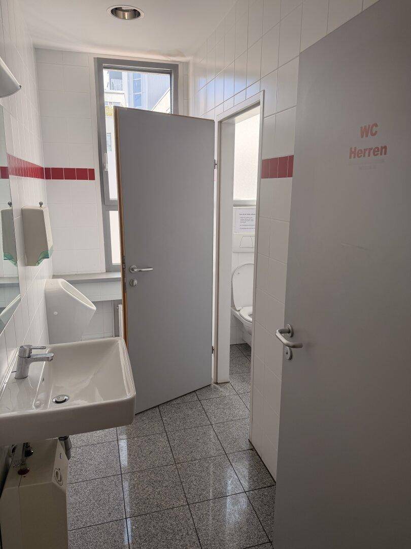 Herren-WC mit Urinal, Waschbecken und Fenster, ausgestattet mit Fliesenboden und weißen Wänden.
