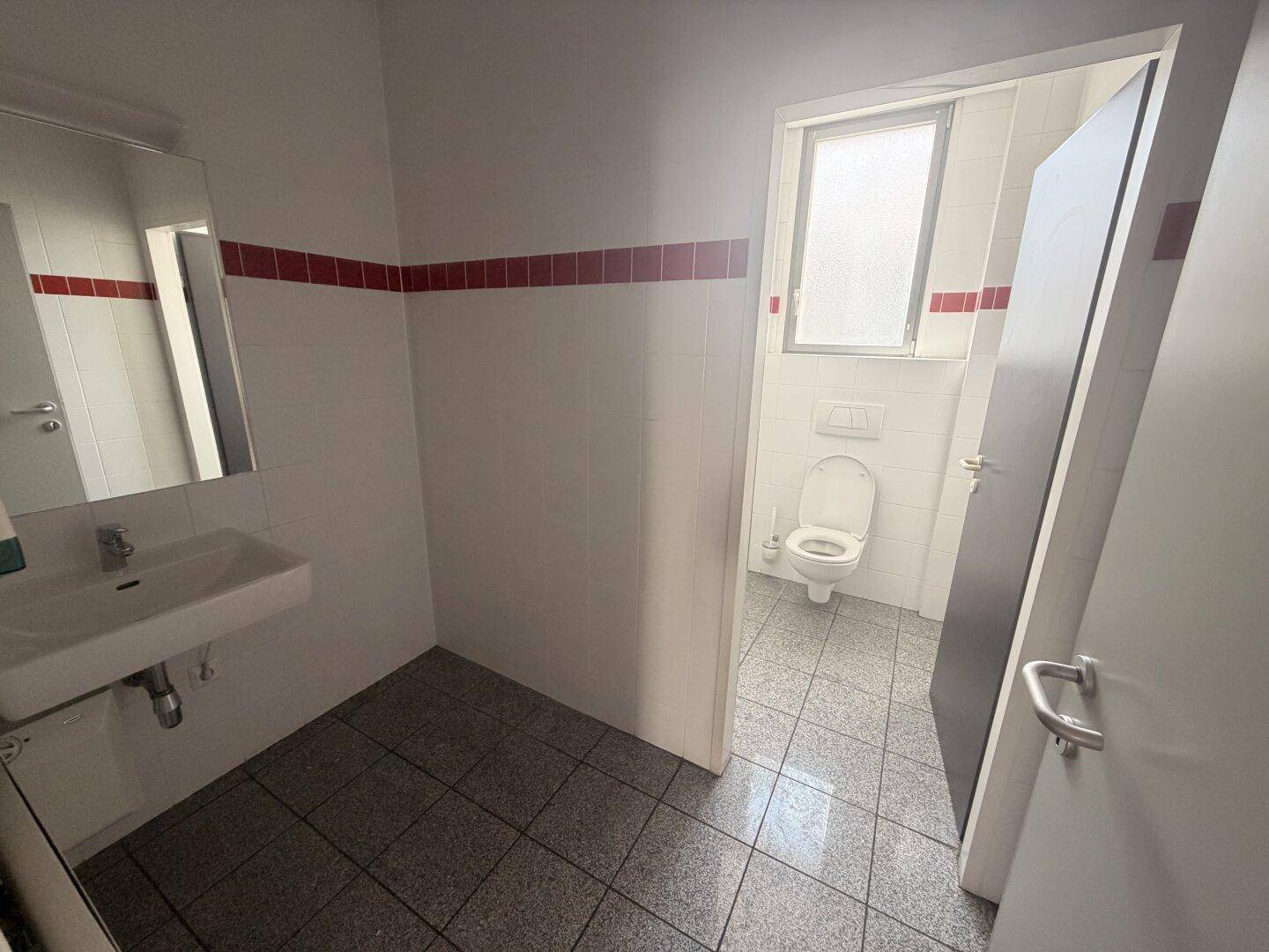 Toilettenbereich mit Waschbecken, Spiegel und Fenster, gefliestem Boden und weißen Wänden.
