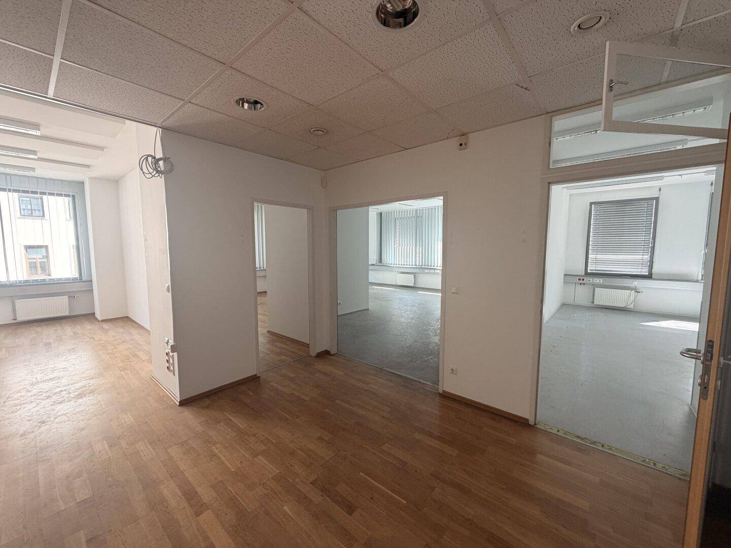 Mehrere leere Büroräume mit Holzparkett und Teppichboden, durch Türen verbunden und mit Jalousien an den Fenstern.