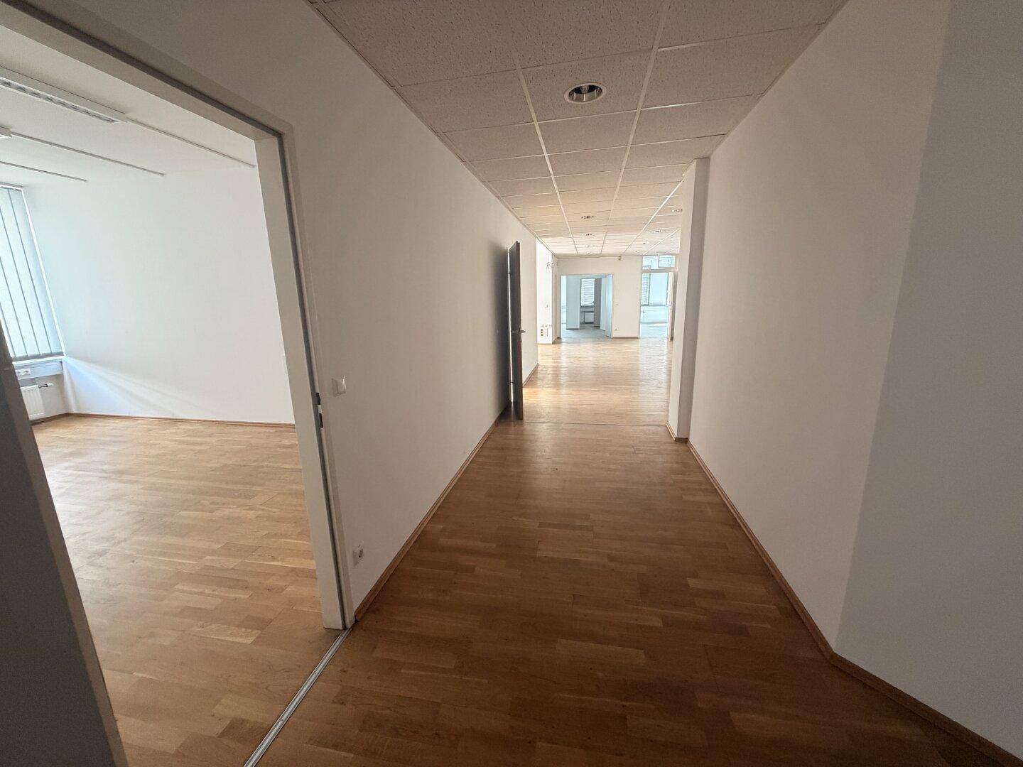 Langer Flur mit Holzparkettboden und weißen Wänden, der zu verschiedenen Büroräumen führt.