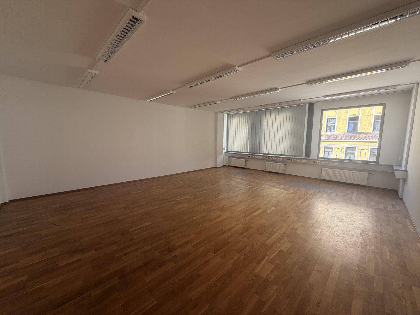 Geräumiger Büroraum mit Holzparkett und breiten Fenstern, die einen Blick auf die Stadt bieten.