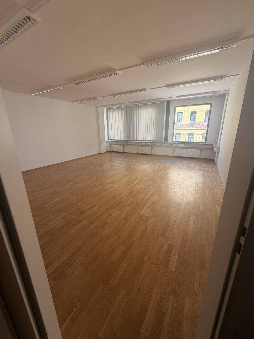 Heller Büroraum mit Holzparkett und großen Fenstern, die viel Tageslicht hereinlassen und eine Stadtansicht bieten.