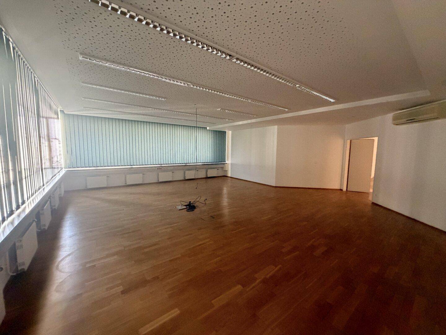 Großer Büroraum mit Holzparkett und durchgehenden Fensterfronten, ausgestattet mit Jalousien und Klimaanlage.