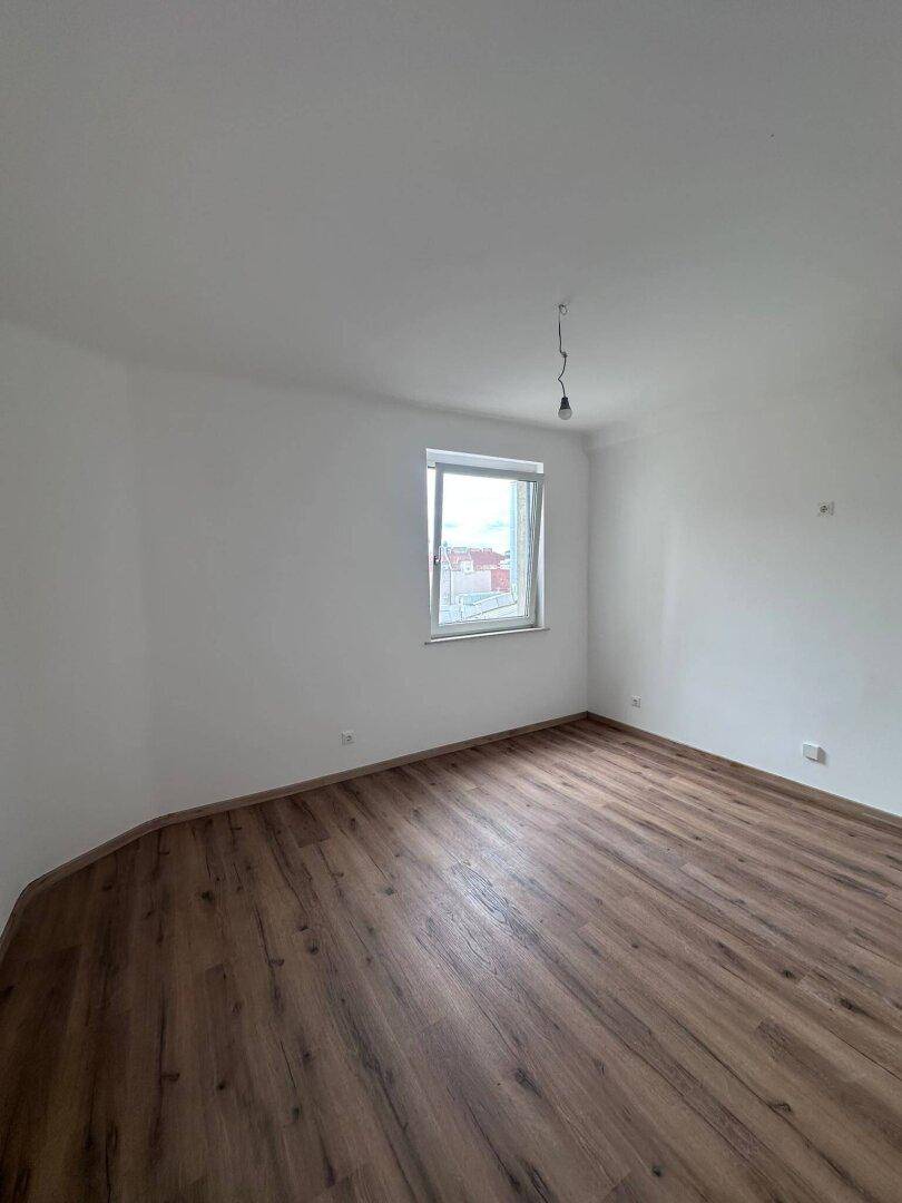 Heller, leerer Raum mit Laminatboden und einem Fenster, das Tageslicht hereinlässt.