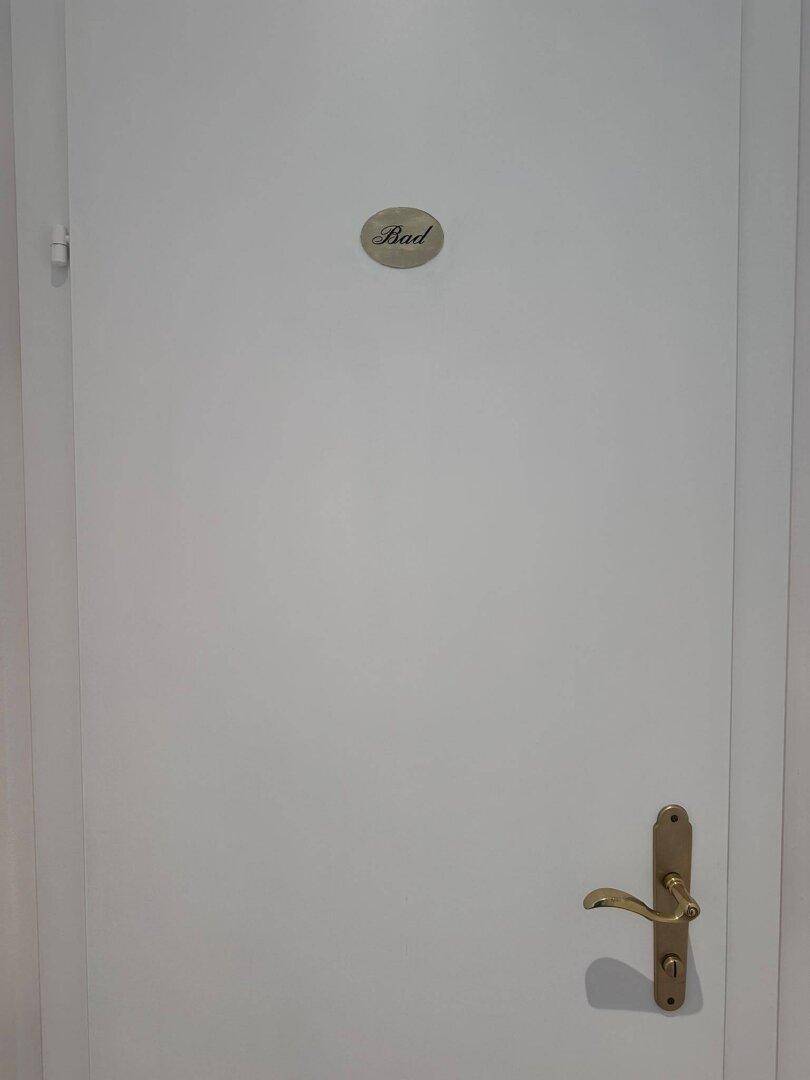 Weiße Tür mit goldener Klinke und einem Schild, das auf das Badezimmer hinweist.