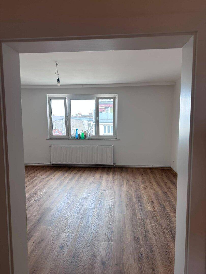 Blick durch eine Tür in ein helles Wohnzimmer mit Laminatboden und Fenster.