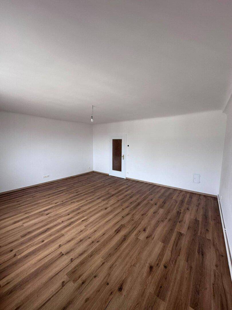 Heller Wohnraum mit Laminatboden und einer Tür, die zu einem anderen Bereich führt.