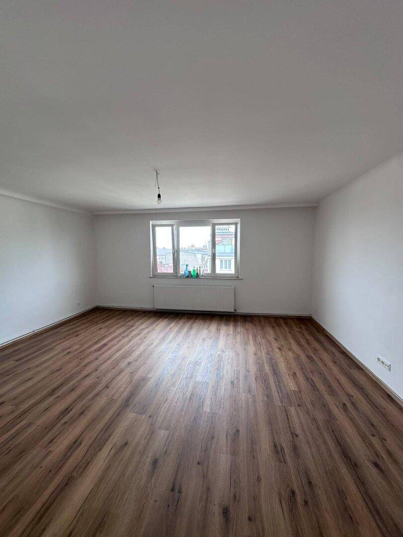 Großes Wohnzimmer mit Laminatboden und breitem Fenster, das den Raum hell erscheinen lässt.