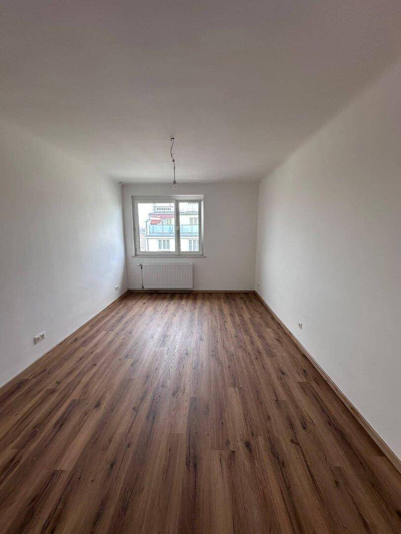 Langes, helles Schlafzimmer mit Laminatboden und einem Fenster, das Tageslicht hereinlässt.