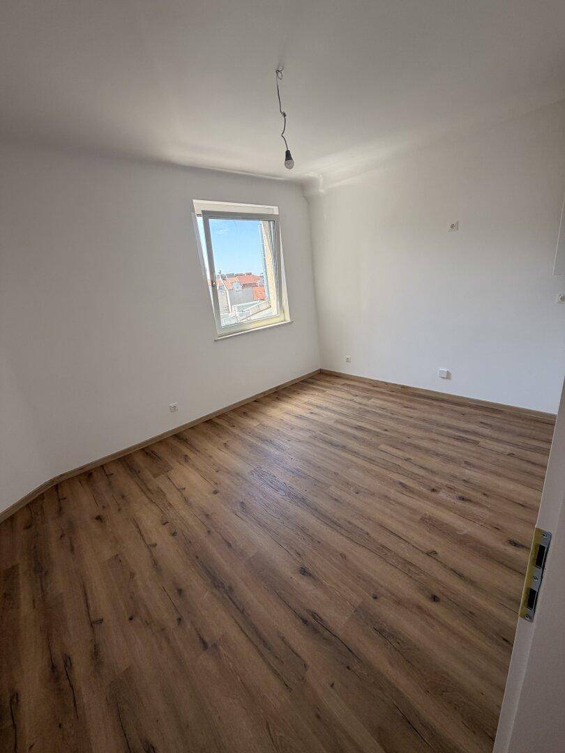 Helles, leeres Zimmer mit Laminatboden und einem Fenster, das viel Tageslicht hereinlässt.