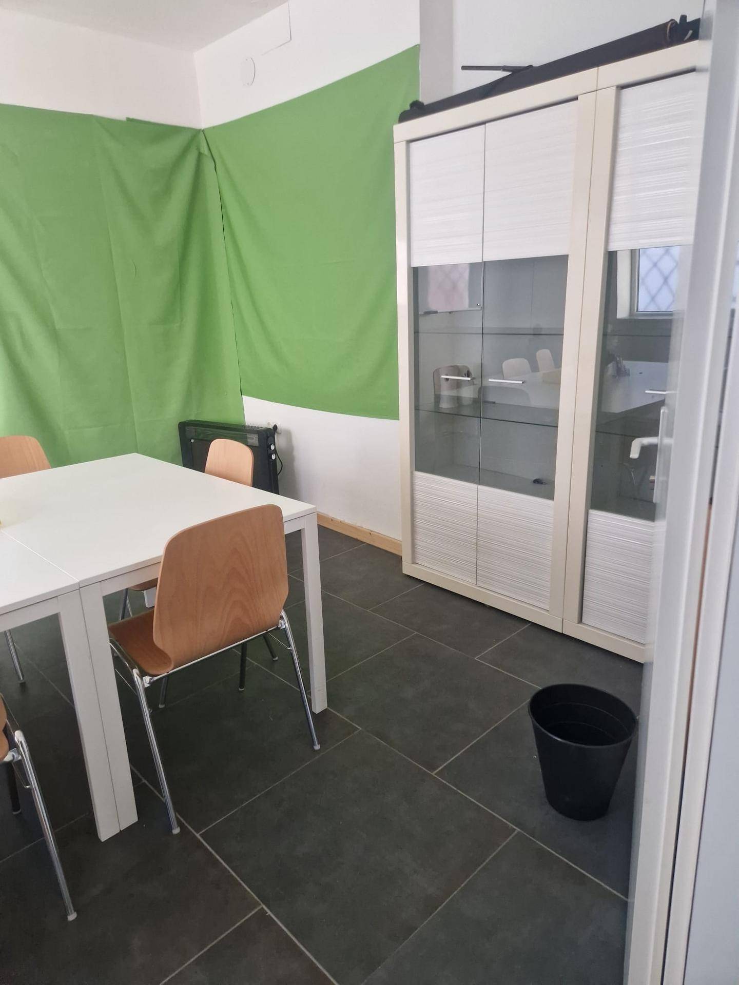 Besprechungsraum mit einem großen weißen Tisch, Holzstühlen und einem grünen Hintergrundtuch. Der Boden ist gefliest.