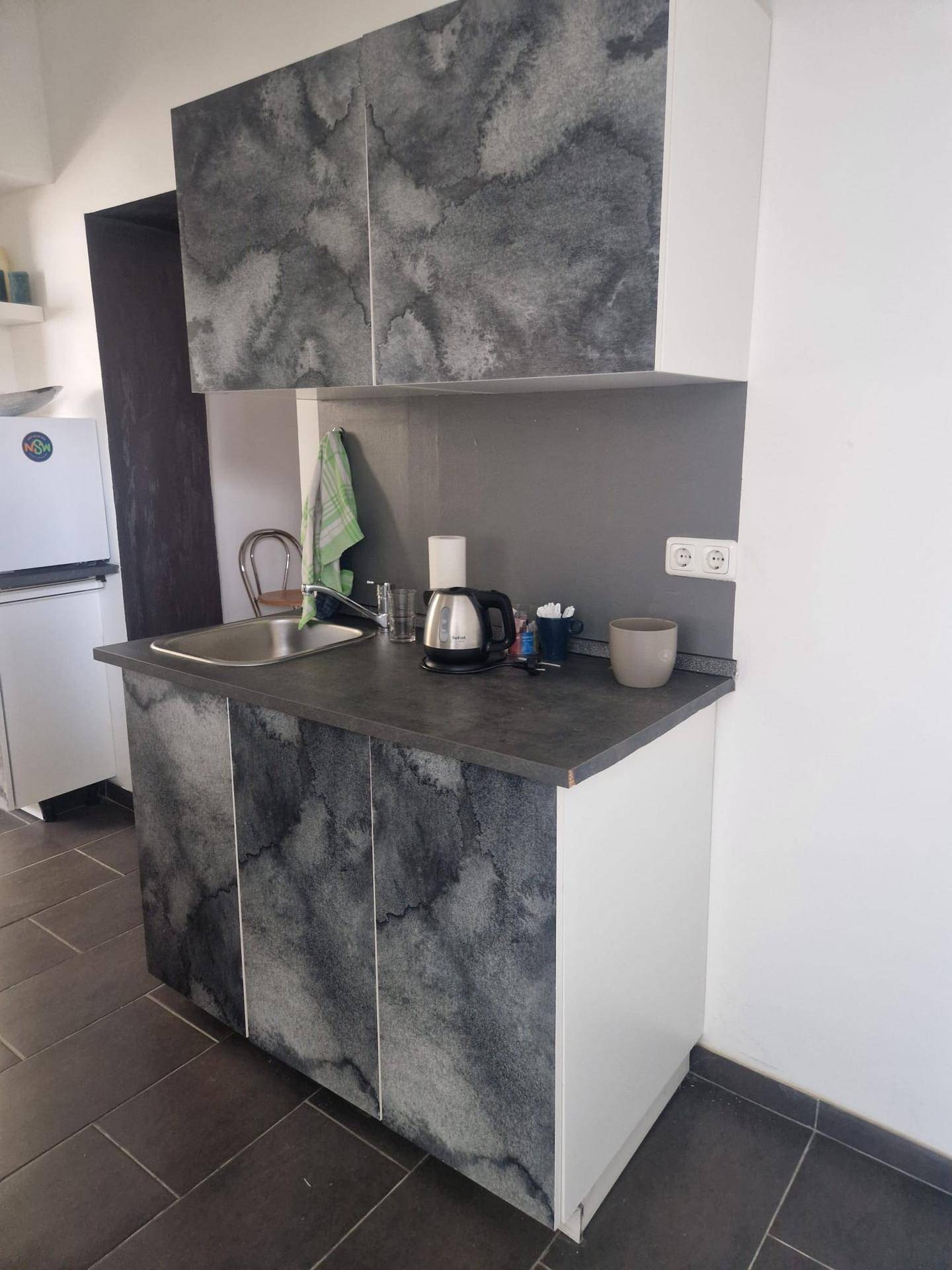 Kleine Küchenzeile mit Spüle, Wasserkocher und Kühlschrank. Die Schränke haben eine graue Marmoroptik.