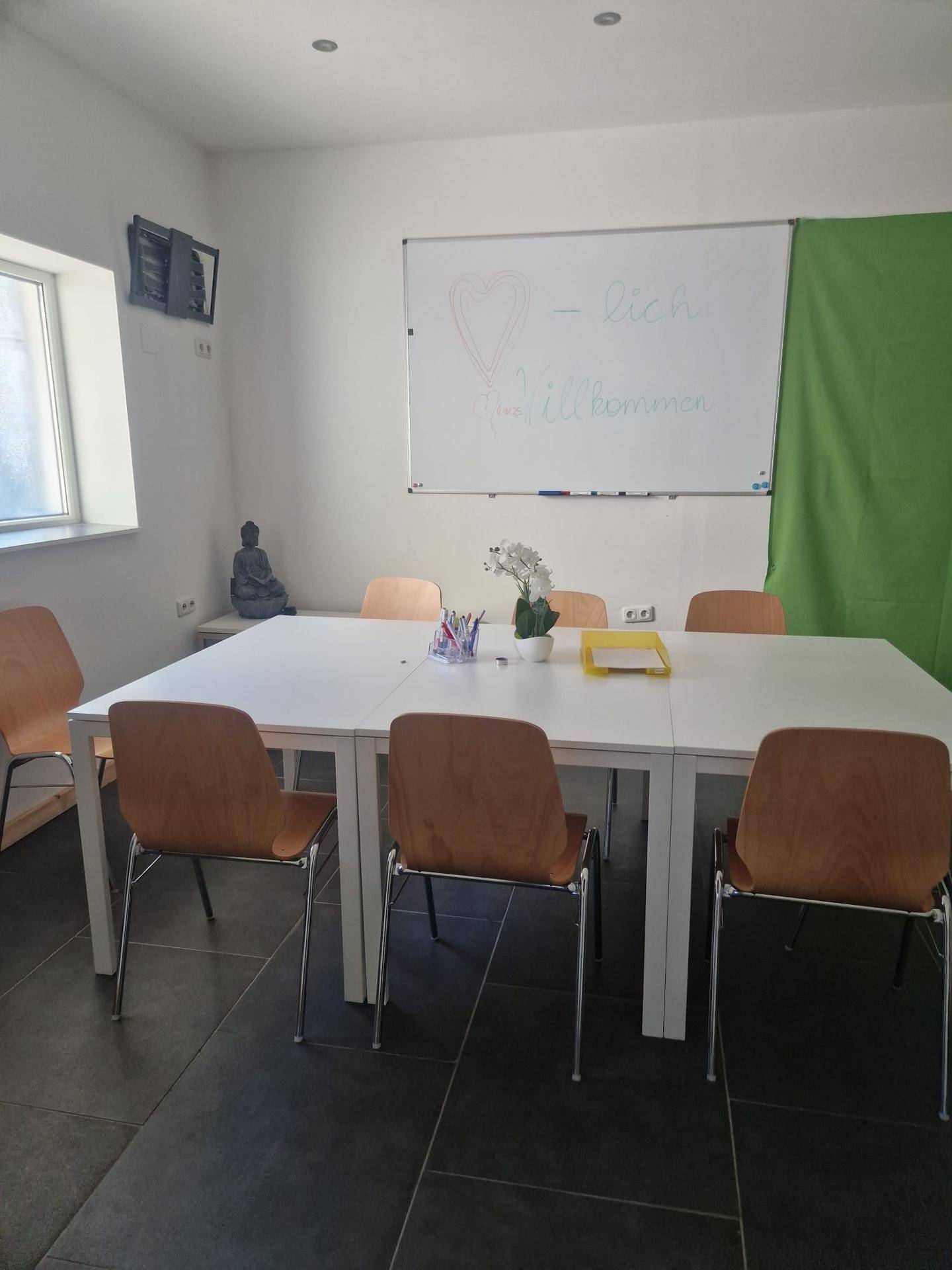 Heller Besprechungsraum mit einem großen Tisch, Stühlen und einem Whiteboard. Ein Fenster sorgt für natürliches Licht.