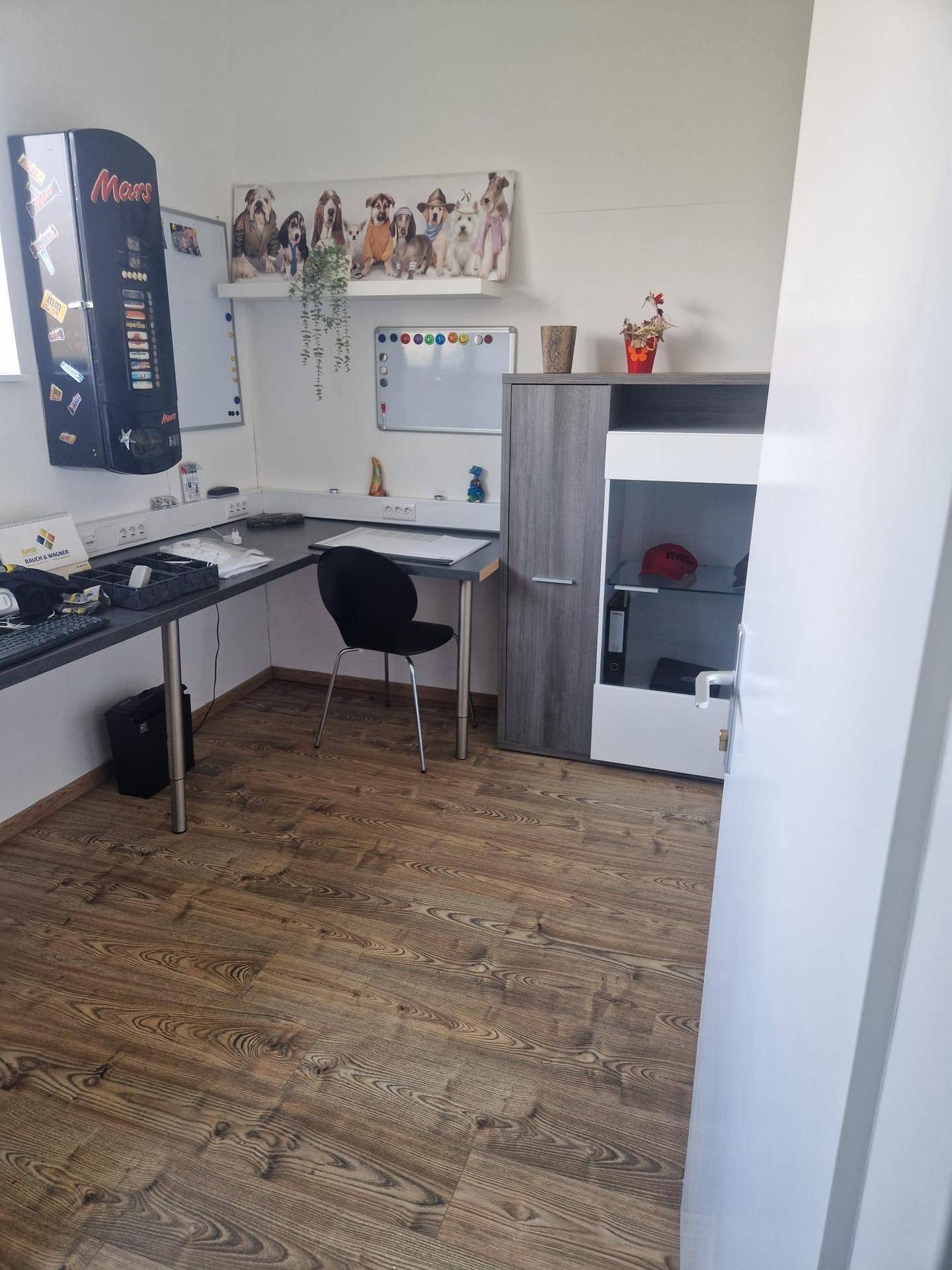 Büro mit einem Schreibtisch, einem Aktenschrank und einem Verkaufsautomaten. Der Boden ist mit Laminat ausgelegt.