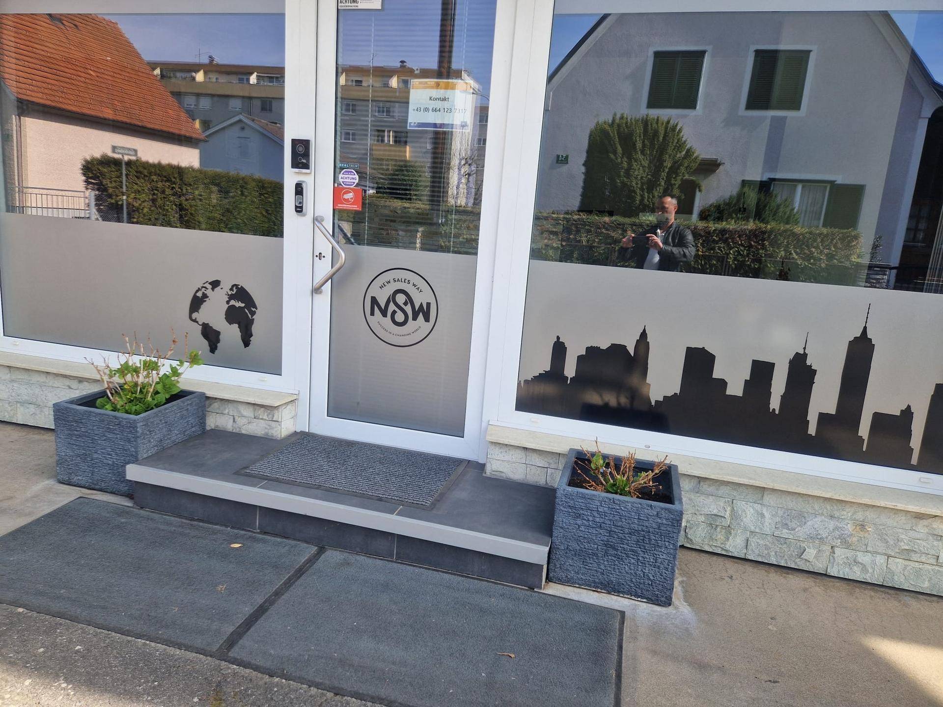 Nahaufnahme des Eingangsbereichs eines Geschäfts mit großen Fenstern und Werbeelementen. Die Steinverkleidung der Fassade ist gut sichtbar.