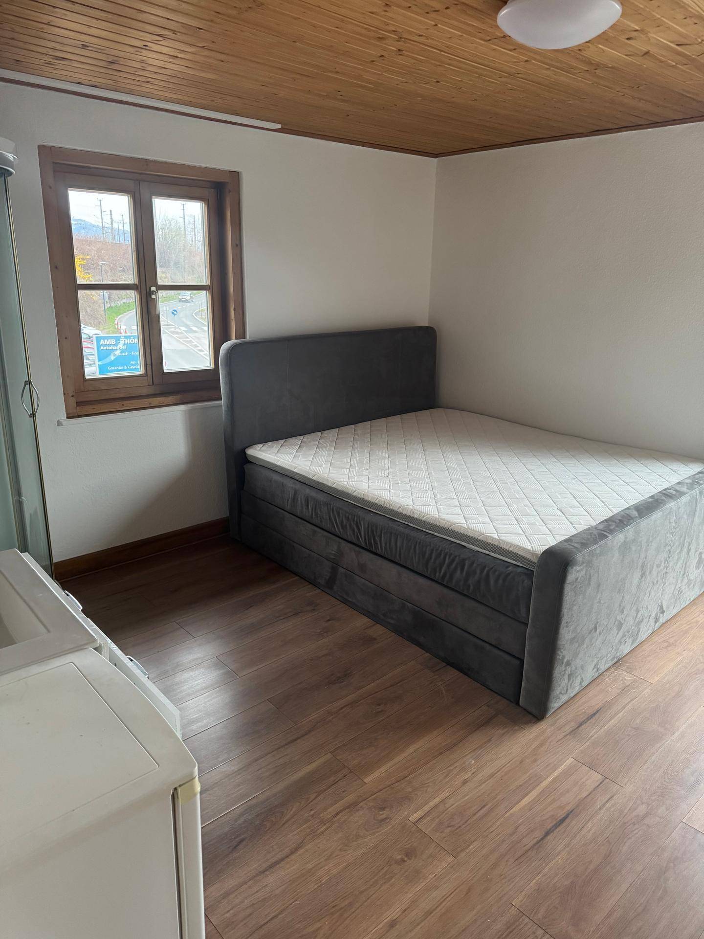 Schlafzimmer mit Holzboden und -decke, einem Bettgestell und einem Fenster mit Blick ins Grüne.