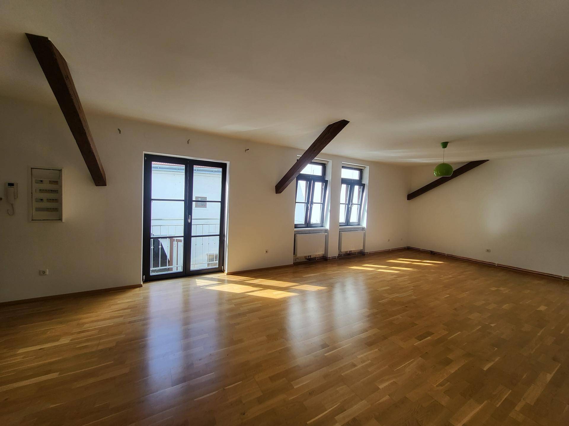 Heller Wohnraum mit Holzbalken, Parkettboden und einer Balkontür, die viel Sonnenlicht hereinlässt.