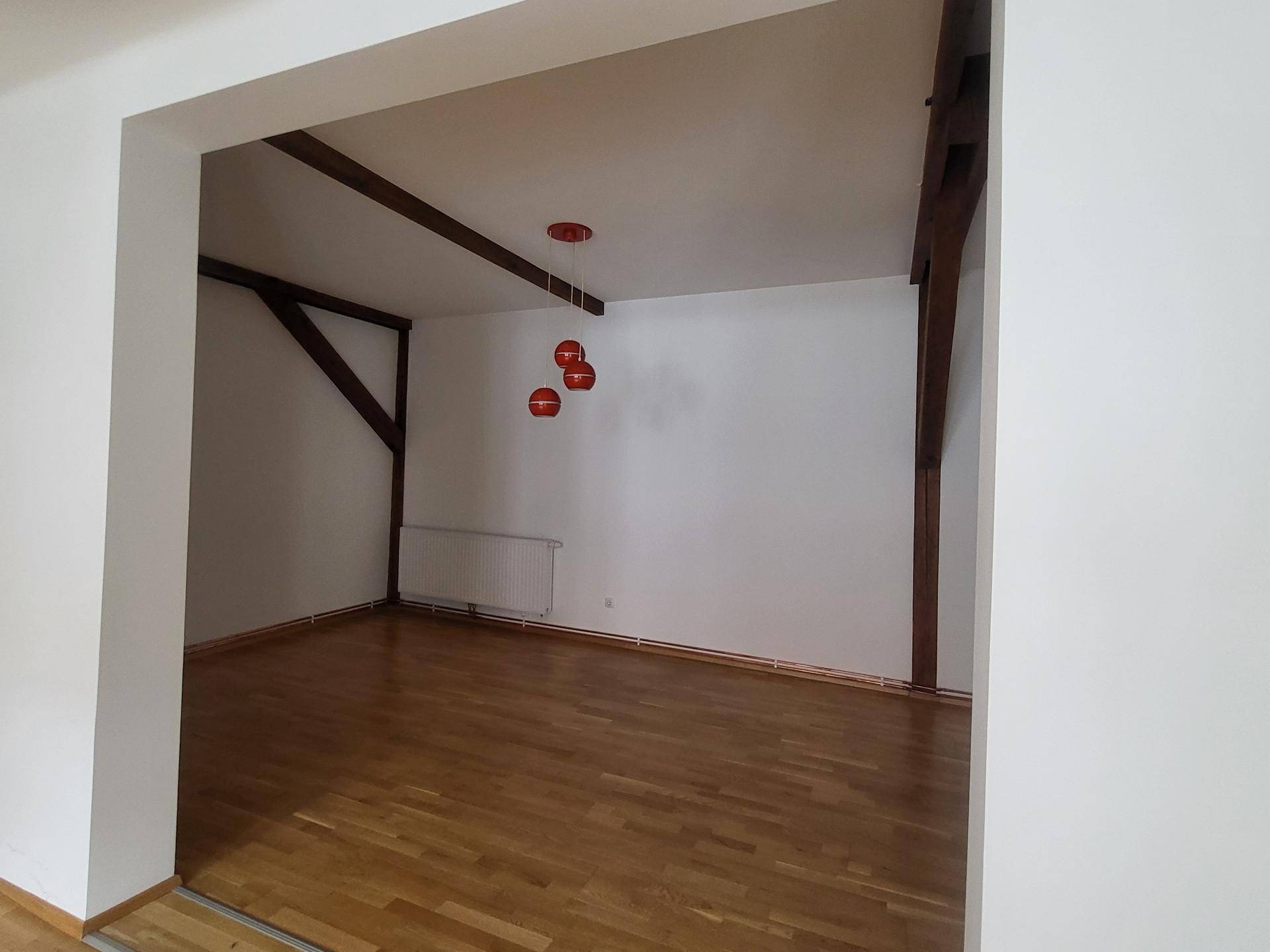 Geräumiger Wohnbereich mit sichtbaren Holzbalken und Parkettboden, der durch einen breiten Durchgang zugänglich ist.
