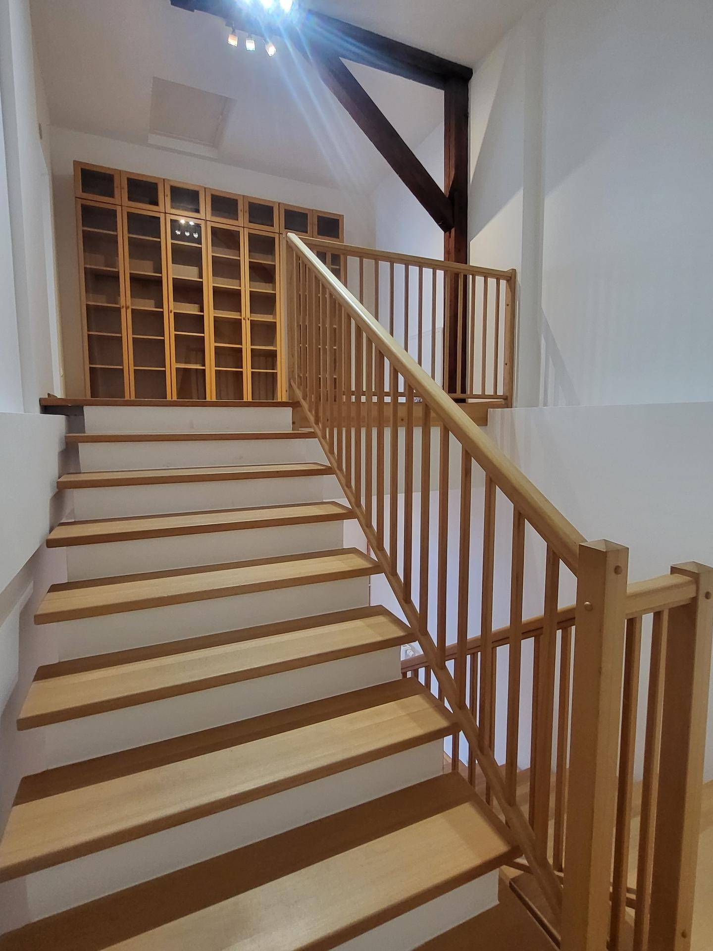 Holztreppe mit Geländer und eingebautem Regal im oberen Bereich, beleuchtet von Deckenstrahlern.