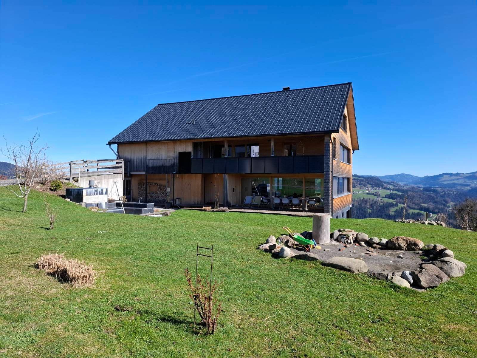Gesamtansicht des modernen Holzhauses mit großem Garten, Feuerstelle und Bergblick.