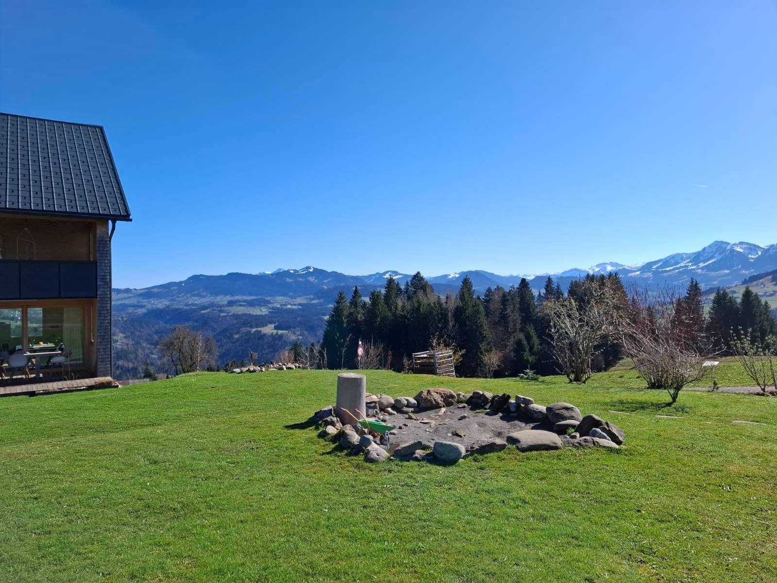 Garten mit Feuerstelle und weitem Blick auf die Berge und grüne Wiesen unter blauem Himmel.