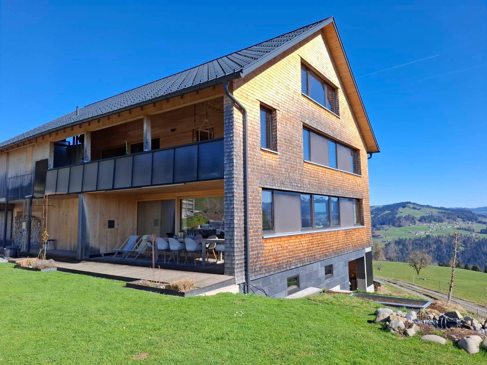 Ansicht des modernen Holzhauses mit großer Terrasse und Balkon, eingebettet in eine grüne Hügellandschaft.