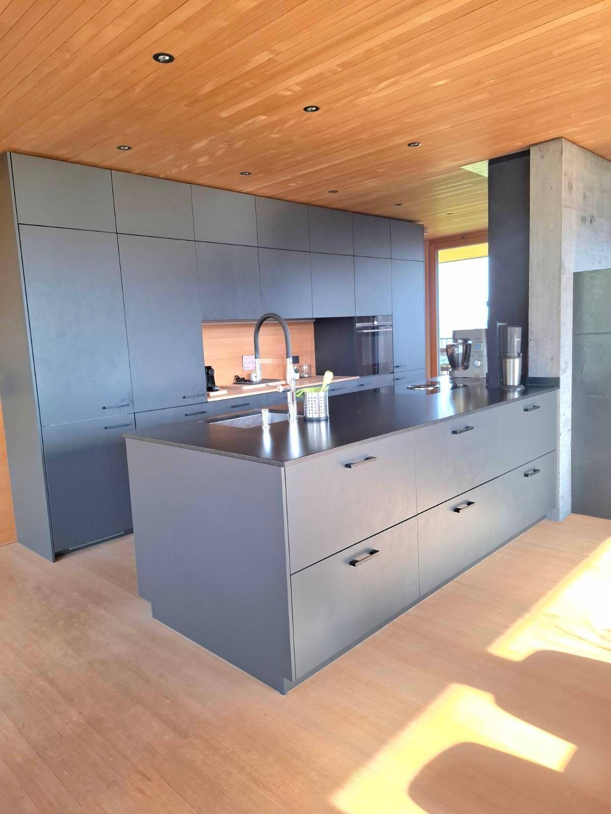 Geräumige, moderne Küche mit Kücheninsel, dunklen Fronten und Holzboden in hellem Ambiente.