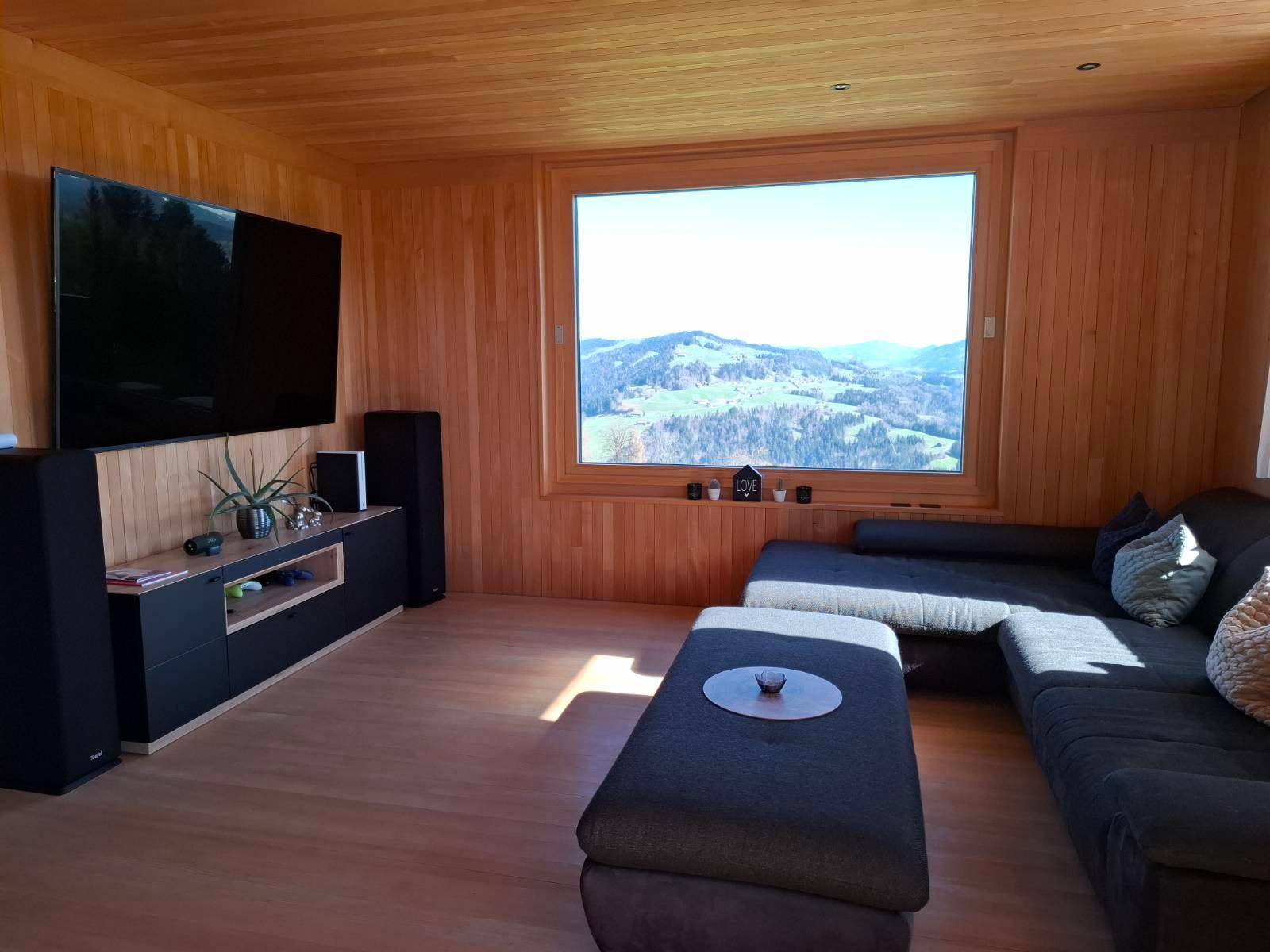 Gemütliches Wohnzimmer mit Holzwänden, großem Sofa und weitem Blick auf die umliegende Berglandschaft.