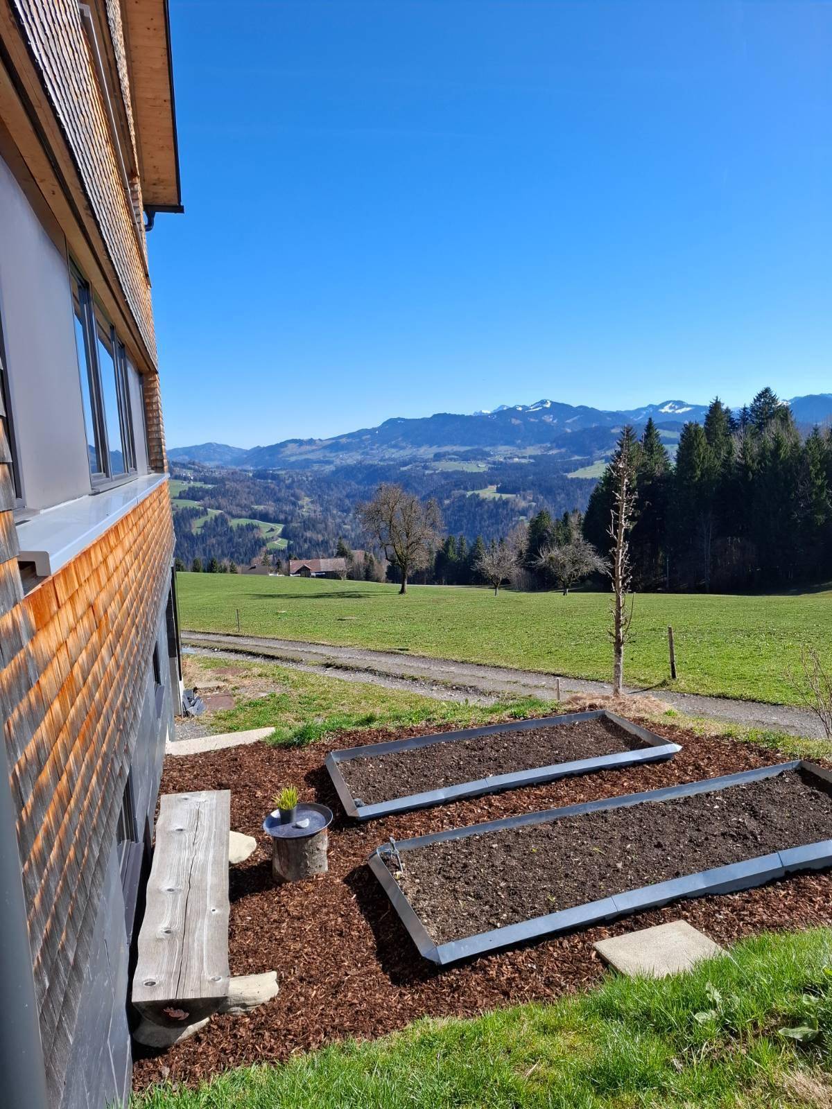 Gartenbereich mit Hochbeeten und einer Holzbank, umgeben von grünen Wiesen und Bergblick.